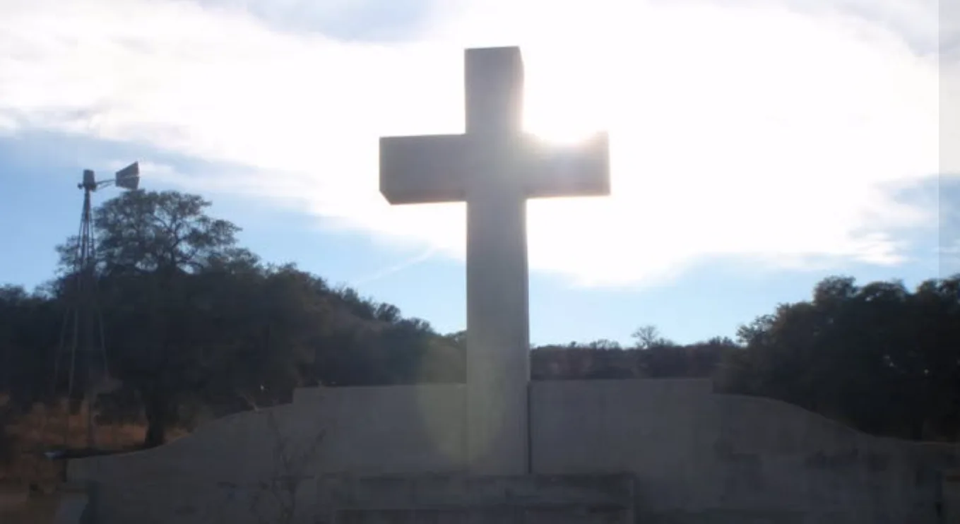 A stone cross backlit by the sun against a bright sky, with a windmill and trees in the distance